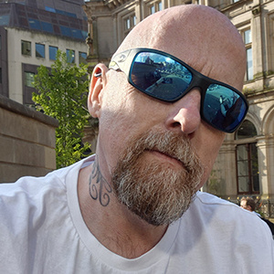 A headshot of Dave McNamara, a man with a bald head wearing a white t-shirt and looking at the camera.