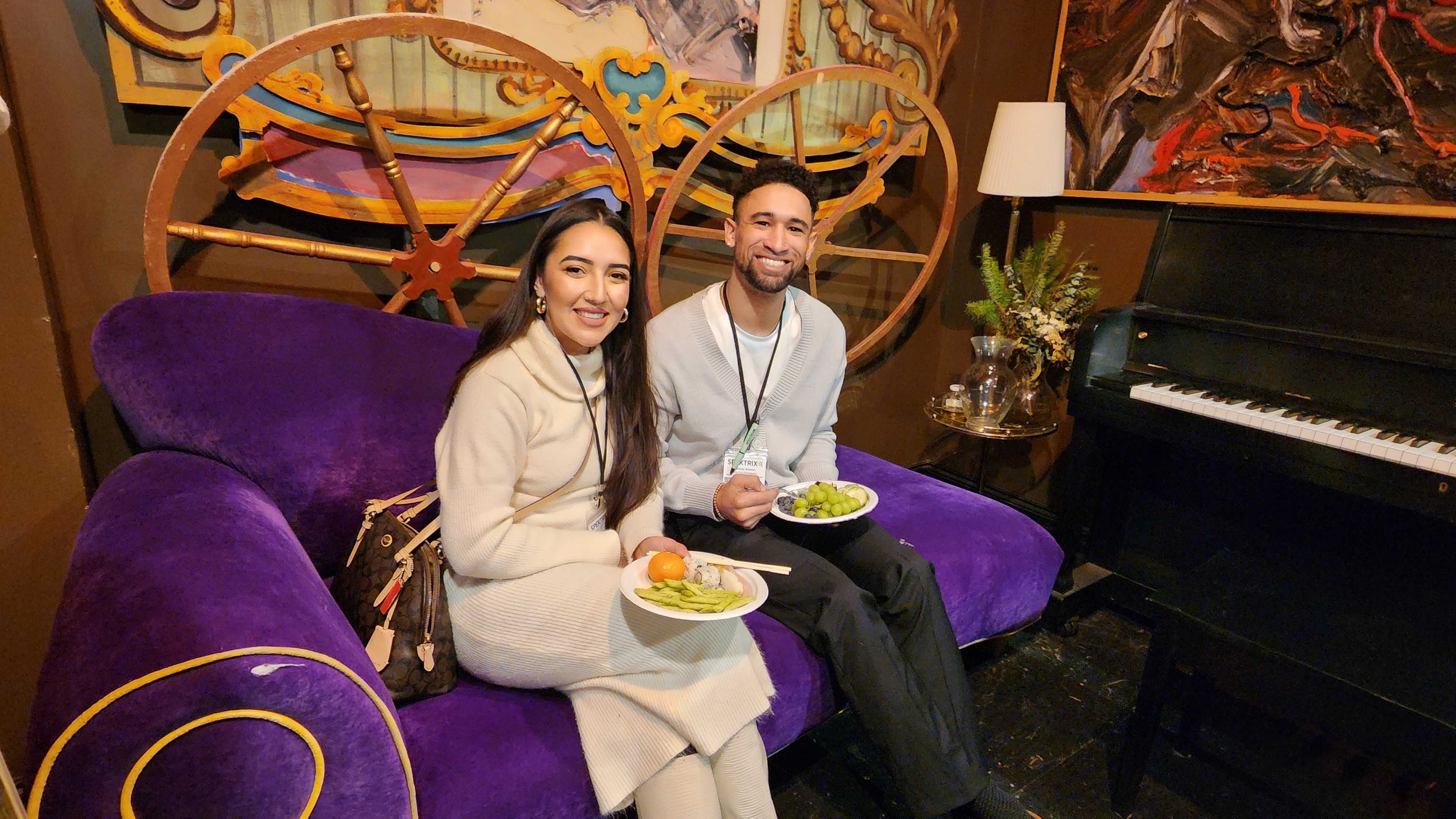 Two members of Santa Cruz Shakespeare sit on a purple couch with plates of food, smiling