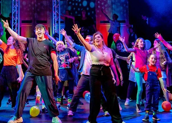 Young performers dance and sing onstage at the Lawrence Batley Theatre, wearing bright colours and surrounded by balloons