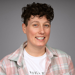 Jade, a white woman, with dark brown curly short hair, wearing a light checked shirt and white t-shirt, looking at the camera smiling.