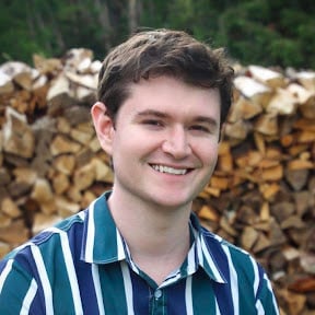 Jake, a white man in his 20s with brown hair, wearing a striped blue, green, and white button up shirt, smiling at the camera in front of a forested background.