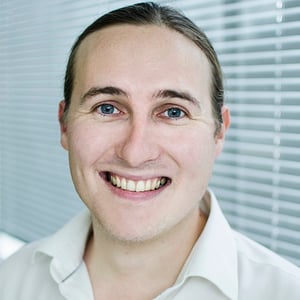 A headshot of Martin Counter, a man in his thirties with long brown hair tied back in a pony tail.  He is wearing a white shirt, standing in front of some window blinds and smiling at the camera