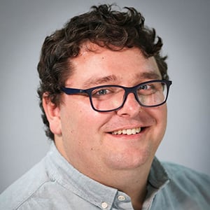 A headshot of Stephen Cripps, a man with dark wavy hair, wearing a blue shirt and glasses smiling