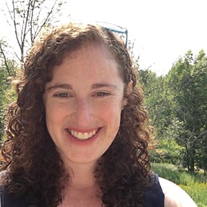 Kate Herzlin, a woman with red curly hair, wearing a dark blue shirt and smiling against a landscape of trees.