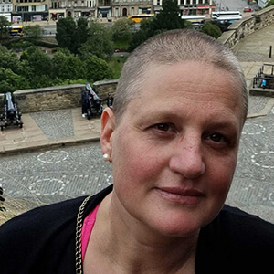 Liv Nilssen, a white woman in her 40s with very short brown and silver hair. In the background a view from Edinburgh Castle showing other visitors, gardens, the street and shops. (1)