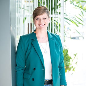 A woman with short light brown hair, wearing a white shirt and green blazer, smiling into the camera, pictured with palm leaves in the background.