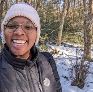 Shanice Brandon, a Black woman in her late 20's, smiles in front of the camera, wearing a light pink winter hat and a black puffer jacket