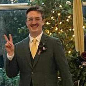 Chris, wearing a forest green suit with a yellow tie and clear-framed glasses, waves the peace sign while smiling into the camera.